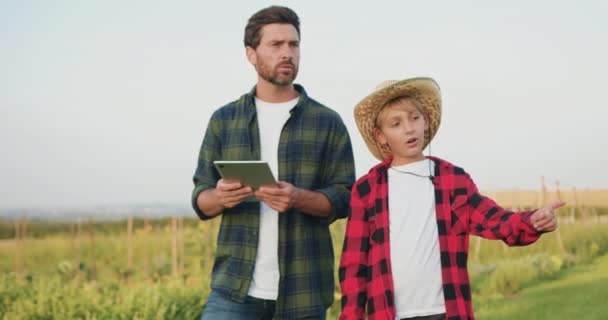 農民労働者男性と10代の息子は フィールドで働くタブレットで帽子を被り 日没時に熟した小麦を収穫するハーベスターを組み合わせます 労働者は小麦の収穫をクリップボードで調べる 小麦生産について — ストック動画