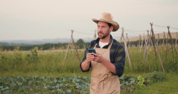 エプロンとゴムブーツの男は スマートフォンでノートを取ります トウモロコシ畑を歩いているシャベルと電話を持つ農民 田舎の道を歩く犬と一緒にゴムブーツの農民 ライフスタイル農家検査 — ストック動画