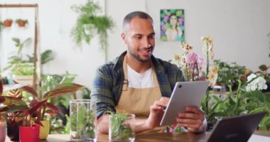 Afro-Amerikan bir adamın portresi dijital tablet bilgisayar kullanıyor ahşap masada botanik bitkilerle oturuyor ve gülümseyen kameraya bakıyor. Çiçekçi dükkanında çalışan erkek satıcı