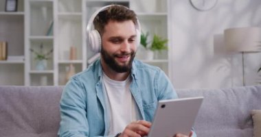 Kulaklıkla gülümseyen beyaz adam dijital tablet kullanarak e-öğrenme, faturaları internet üzerinden ödeme, e-ticaret, uzaktan çalışma, evde sosyal medya yapıyor. Yavaş çekim