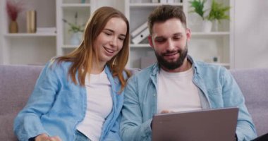 30 'lu yaşlardaki genç bir çiftin portresi kanepeye oturup tablet kullanıyor. Genç adam, kız arkadaşına yeni mobil uygulamalar ve hafta sonları modern teknoloji kullanımını açıklıyor. Yavaş çekim.