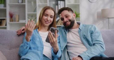 Kocasına akıllı telefon kullanarak sosyal ağda video içeriği gösteren gülümseyen genç bir kadın. Mutlu aile çifti, modern teknoloji aletleriyle evdeki koltukta birlikte oturuyorlar. Yavaş çekim