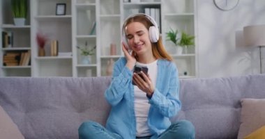20 'li yaşlarda, kulaklıklı genç bir kadın koltukta oturuyor ve cep telefonu kullanıyor. Yan tarafa bakıyor, müzik dinliyor evde dinleniyor, boş vakitlerini içeride, oturma odasında geçiriyor.