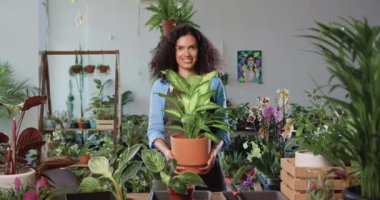 Çiçekçide dikilip gülümseyen ve yeşil saksı bitkisine gülümseyen güzel kadın çiçekçinin portresi. Aile işi ve girişimci kavramı. Yavaş çekim.