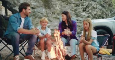 Mutlu gülümseyen aile ve çocuklar hafta sonu doğa pikniğinde ellerini ateşle ısıtıyorlar. Baba, anne ve çocuklar arabanın yanında oturmuş şenlik ateşiyle konuşuyor ve neşeyle iletişim kuruyorlar. Yavaş