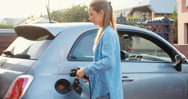 Close-up. Woman plugs in charger into socket of her modern new electric car. Girl in a casual summer outfit, charging a gray electric car parked on the street.