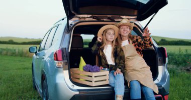 Arabaların bagajında oturan, dijital tablet tutan ve gülümseyen neşeli, modern çiftçi ailesi. Ebeveyn ve çocuk arabada dinleniyor..