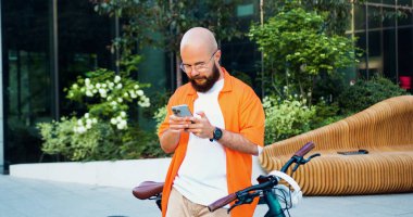 Sabah işe giderken klasik bisikletine binmeden önce akıllı telefon kullanan gözlüklü yakışıklı yetişkin bir adam. Erkek hipster, etkileyici şehir binası geçmişinde telefonla mesaj yazıyor..