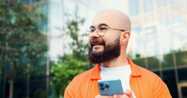 Kapatın. Sakallı adam sokakta cep telefonu ve mesaj kullanıyor. Akıllı telefon kullanırken sosyal medya haberlerini kaydıran gözlüklü yakışıklı adam.