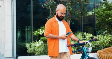 Gündelik giysiler ve gözlük takan beyaz sakallı bir adam şehir merkezinde bisikletle yürürken akıllı telefon kullanıyor. Şehirde sürdürülebilir iş yaşam tarzı.