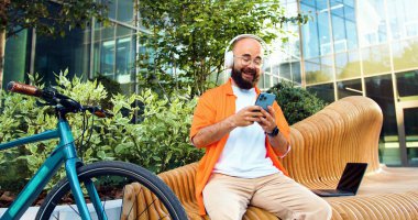 Gözlüklü ve kulaklıklı neşeli hippi adam, akıllı telefon kullanıyor, sosyal ağlarda yazıyor ve internetten çılgınlar gibi sohbet ediyor, arka planda bisikletinin yanında oturuyor..