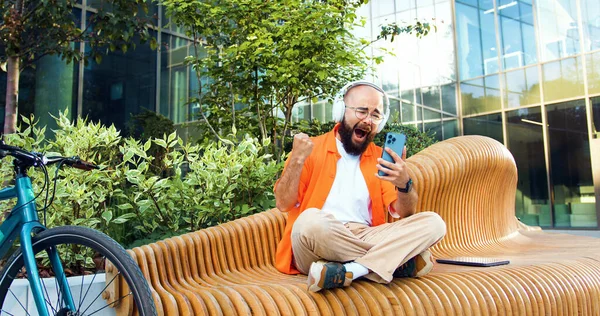 Man in glasses holding phone celebrate good mobile news surprised at work with modern office building. Bearded male person using smartphone and rejoicing while having good news.