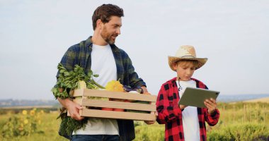 Erkek çiftçi oğluyla sebze tarlasında tablet bilgisayar kullanarak iletişim kuruyor. Adam elinde taze sebze dolu bir kutu tutuyor ve oğluyla gün batımında konuşuyor. Küçük aile şirketi konsepti.