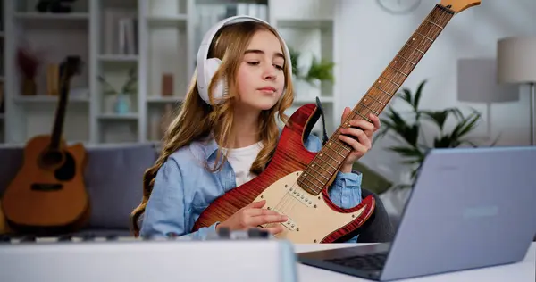 Beautiful teenager girl in earphones learning play guitar at home using online lessons on laptop. Beautiful girl with long hair play e guitar at home for online audience on laptop