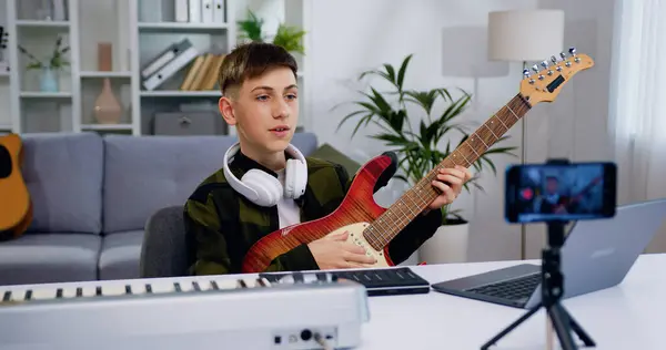 Handsome teenager blogger recording video class tutorial to play guitar on modern smartphone at home studio. Teen boy showing how play for electric guitar for internet auditory.