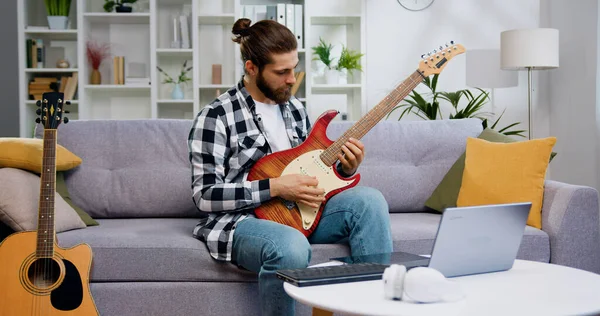 Talented musician conducts class on laptop sitting on sofa in modern studio playing guitar. Caucasian male musician uses musical instrument and conducts video lesson through laptop for students