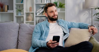Evdeki modern oturma odasında akıllı telefon kullanan mutlu, yakışıklı 30 yaşında bir adam. Kafkasyalı adam internette gezinirken kanepede dinleniyor ve sosyal ağlardaki, evlerdeki videolara bakıyor.