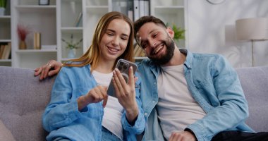 Kocasına akıllı telefon kullanarak sosyal ağda video içeriği gösteren gülümseyen genç bir kadın. Mutlu aile, modern teknoloji aletleriyle evdeki koltukta oturuyor.