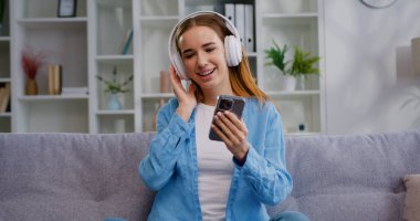 20 'li yaşlarda, kulaklıklı genç bir kadın koltukta oturuyor ve cep telefonu kullanıyor. Yan tarafa bakıyor, müzik dinliyor evde dinleniyor, boş vakitlerini içeride, oturma odasında geçiriyor.