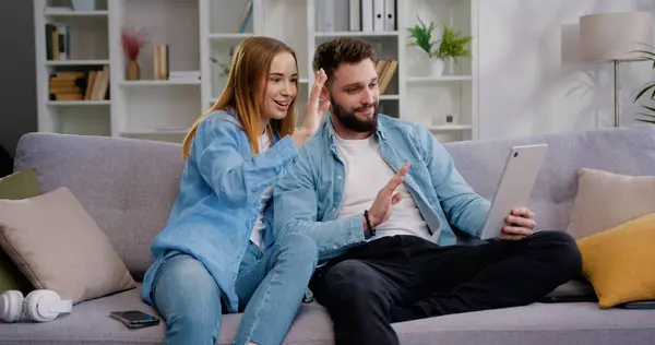Cheerful Caucasian couple man and woman bloggers sitting on couch talking video call smiling waving hello communicate at online conference with relatives recording video blog at cozy living room