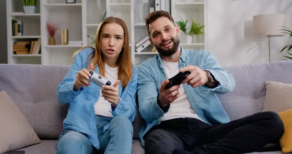 Happy family sitting on couch and playing video games. Adult man and woman uses PlayStation holding console. Games at home together, man and woman, happy family