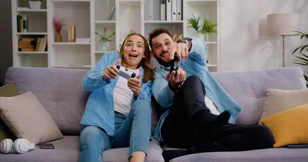 Happy couple sitting on the sofa playing video games, using controllers. competitive Girlfriend and boyfriend in love have fun playing in online video games in living room at home together