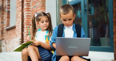 Okulun küçük öğrencileri dışarıda dizüstü bilgisayar kullanıyor. Çocuklar merdivenlerde oturuyor ve dışarıda okul kampüsünün yakınında dizüstü bilgisayarda oynuyorlar. Çeşitli çocuklar tablet ve defterle bir proje hazırlıyorlar..