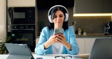 Modern, mutlu ve rahat bir kadın cep telefonuyla cep telefonuyla video izliyor ve favori müzik podcastını veya internet sosyal medyasını dinliyor..