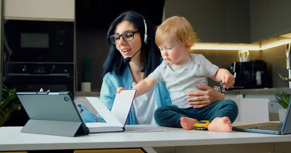 Anne, erkek bebekle uzaktan çalışıyor. Uzak iş konsepti. Kadın evde, mutfakta çalışmaya çalışıyor, bebek çocuk masada oturuyor..