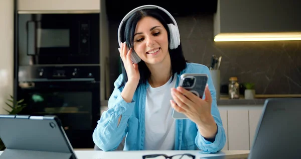 Happy relaxed woman freelancer dancing sitting at laptop wearing headphones listening music using smartphone in modern kitchen at home.