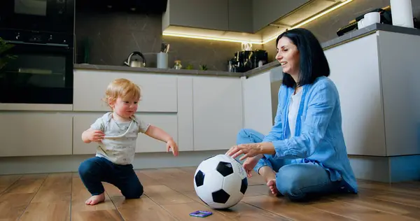 Happy relaxed woman sitting on carpet on the floor at home and smiling while her cute baby son playing with ball. Happy young family resting on the carpet in domestic room.
