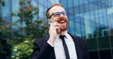 Şehir ticaret bölgesinde cep telefonuyla konuşan bir adamın portresi. Kendine güvenen genç işadamı akıllı telefondan konuşuyor. Gülümsüyor, dışarıda takım elbise giyip mutlu oluyor..