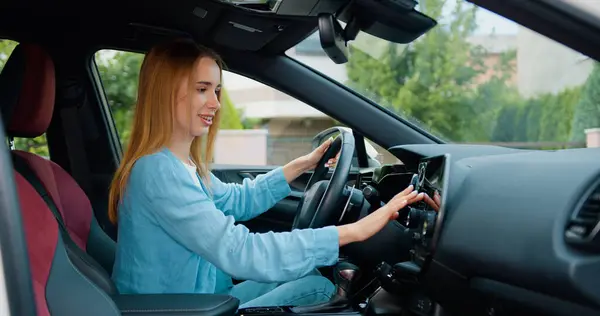 Young woman is sitting in the drivers seat using a touch display screen multimedia device in sporty car driven. Attaractive girl tasting new vehicle functions.