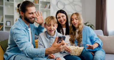 Mutlu aile babaları ve şirin okul çocukları kanepede oturup komik akıllı telefonlar kullanıyor konferans konuşmaları yapıyor, online içeriği izliyor, gülümsüyor, gülüyor..