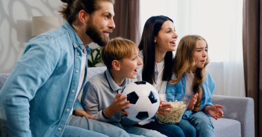Aile evde kanepede oturup futbol maçını canlı yayında izleyerek bir golü kutluyor. Mutlu ebeveynler, anne, baba ve çocuklar top oynayarak en sevdikleri futbolun zaferini destekliyorlar.