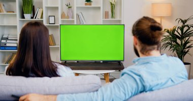 Mutlu çift televizyon izliyor ve kanalları değiştiriyor. Modern oturma odasındaki yeşil ekrana bakıyor. Kadın ve erkek krom televizyon izliyor..
