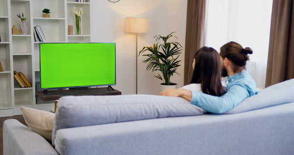 Happy man and woman sitting at home in living room watching green chroma key screen television, relaxing on a sofa. Couple room watching sports game, news, TV show or a movie.
