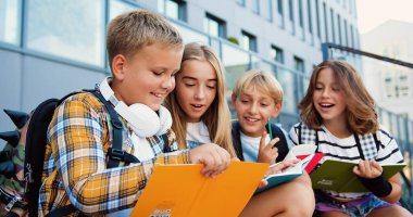 Bir grup ilkokul çocuğu, modern okul binası arka planında açık hava sınıfında kitap yazıyor..