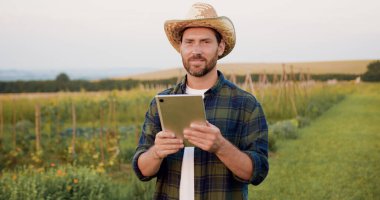 Hasır şapkalı sakallı çiftçi modern teknolojilere erişmek ve tarım alanında ürün üretimini optimize etmek için dijital tablet kullanıyor..