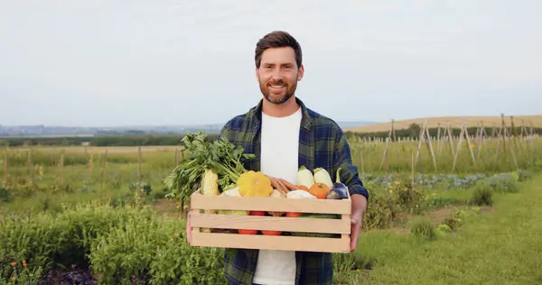 Portrait handsome caucasian farmer holding wooden box of colorful vegetables. Happy Smiling agriculture worker looking at camera and smiling while standing in field harvest garden nutrition organic