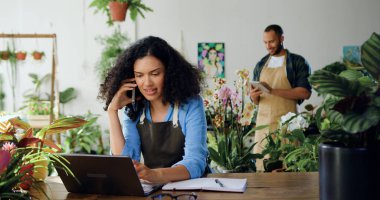 Önlüklü Afro-Amerikalı kadın, müşterisiyle cep telefonuyla konuşuyor ve bilgisayarcı adam kullanıyor. Aile çiçekçileri dizüstü bilgisayar kullanıyor, daktilo ediyor ve konuşuyor.