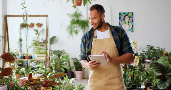 African american male gardener using digital tablet and purchasing flower orders. Man florist checking green plants in flower shop and using computer touching screen. Floristry and modern technology