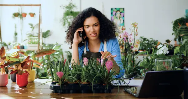 Happy african american woman in apron sitting on desk speaking to client on mobile phone and using laptop looking at flower shop green plants. Female florist in flower store orders flowers from