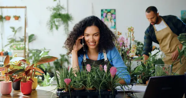 African american female worker of flower shop sitting at table and calling on smartphone and caring plants in planner while sitting in cozy floral shop. Woman florist flower store owner working. Small