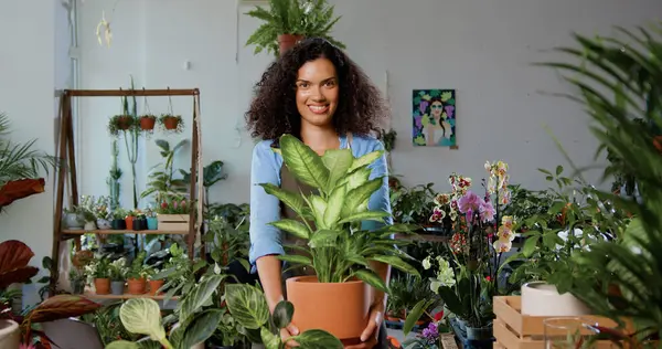 Portrait of good-looking woman florist worker in uniform standing in flower store and smiling showing into camera green seedling pot plant. Concept of family business and entrepreneur.