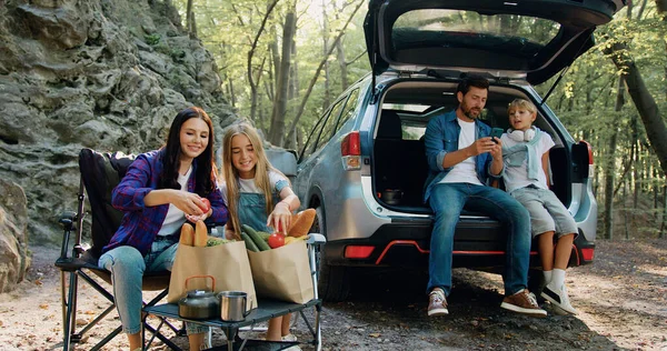 Happy family having weekend picnic outdoors in the forest. Woman and girl preparing food and father and son siting in open trunk of car and using phone. Time to Travel concept. Family travel dream
