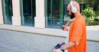 Hippi, şehir merkezinde bisikletle yürürken akıllı telefon kullanıyor. Şehir caddesinde modern yeşil bisikletli, şık giyimli, kendinden emin genç bir erkek portresi..