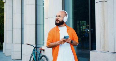 Kulaklıklı genç adam cep telefonu kullanarak mesaj yazıyor ve bisikletle sokakta yürüyor. Meşgul insanlar için modern aletler.