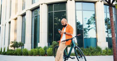 Sabah işe giderken klasik bisikletine binmeden önce akıllı telefon kullanan gözlüklü yakışıklı yetişkin bir adam. Erkek hipster, etkileyici şehir binası geçmişinde telefonla mesaj yazıyor..