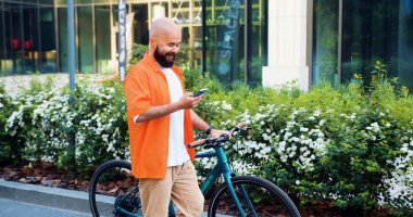 Beyaz adam, modern şehir binasının arka planında bisiklete yaslanırken bir mesaj yazarken akıllı telefon kullanıyor. Kentte bisiklet, kentsel şehirde yürüyen eko taşıma mobil uygulaması.